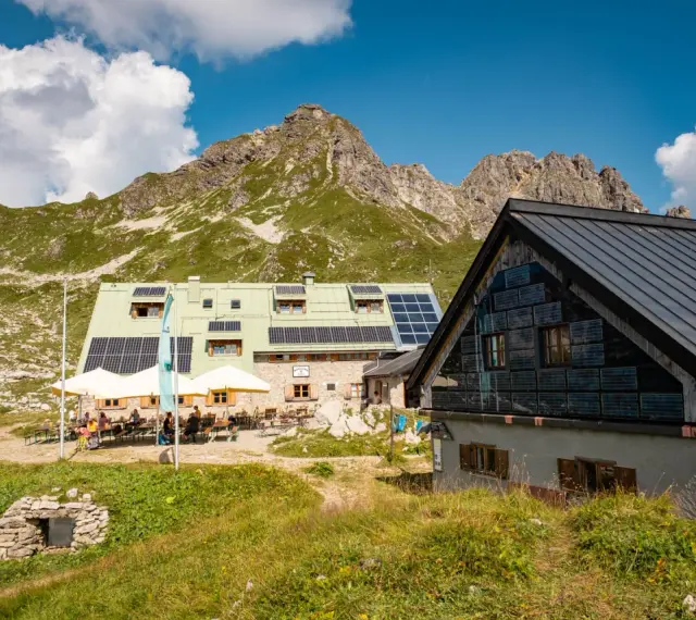 Die Buchel Alpe, eine Berghütte mit Solaranlagen, liegt vor einer beeindruckenden Bergkulisse. Gäste sitzen draußen unter Sonnenschirmen.