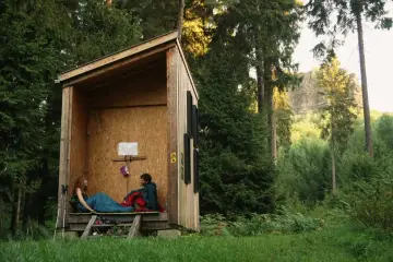 Zwei Personen entspannen sich in einer einfachen Holzhütte, bereit für wild camping. Umgeben von hohen Bäumen und üppigem Grün.