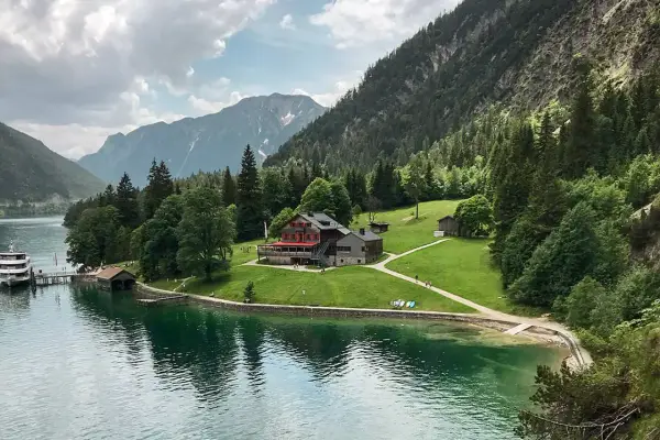 Gaisalm am Achensee: ein Gasthaus mit roten Fensterläden, umgeben von grünen Wiesen und Bergen. Ein Schiff liegt am Steg.