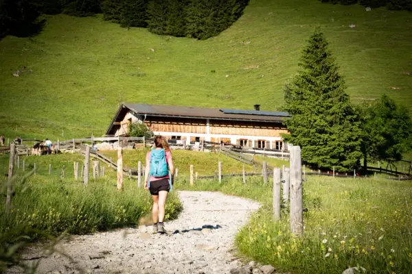 Frau mit Rucksack wandert auf einem Schotterweg zu einer Berghütte. Grüne Wiesen und Bäume im Hintergrund.