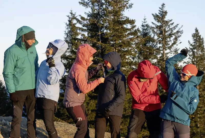Sechs Personen in bunten Hardshelljacken stehen vor Bäumen. Eine Hardshelljacke schützt vor Wind und Wetter.